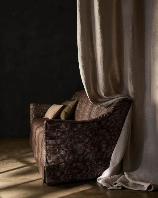 Brown upholstered couch with cushions beside draped beige curtains against a dark grey wall in an indoor setting with subtle lighting.