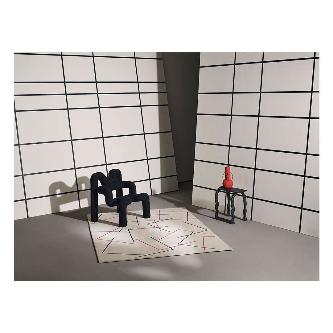 Contemporary interior photo features a black chair, patterned rug, black side table with orange vase against a white-paneled wall.