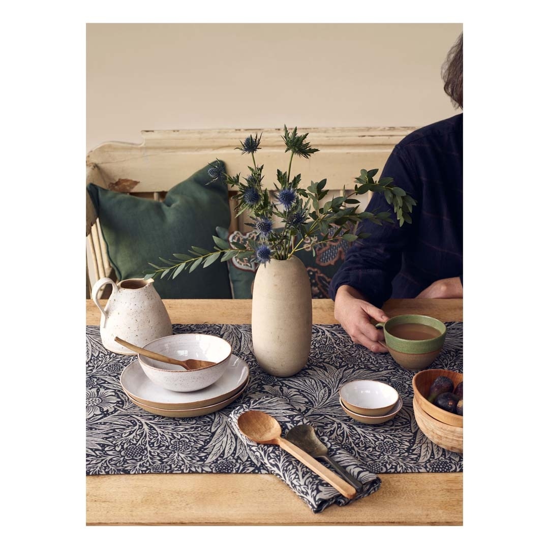 Table setting featuring a person with coffee, vase of thistles, ceramic dishes, wooden utensils, and a patterned table runner indoors.