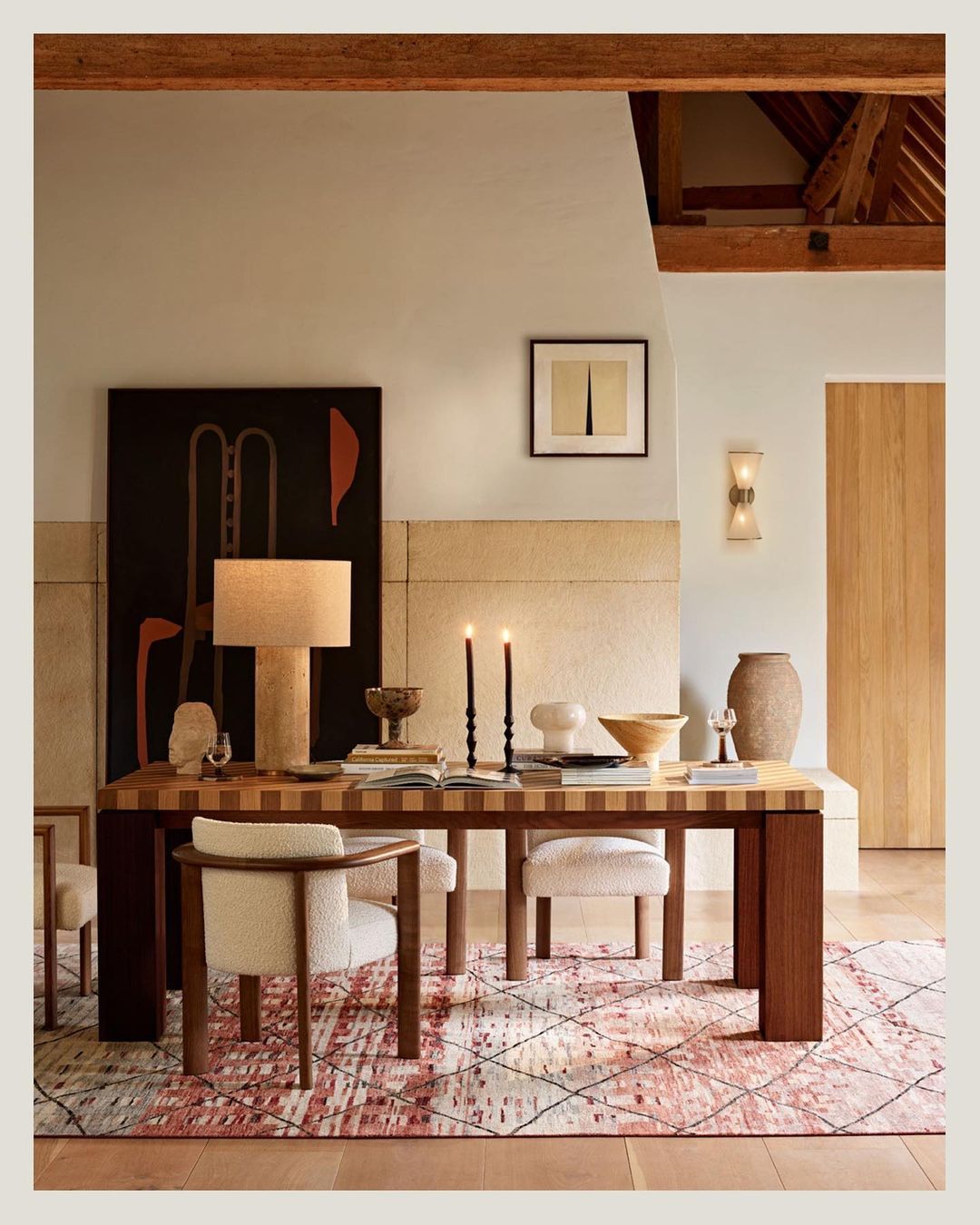 Interior view showcases a wooden table set with books, ceramics, and candles, alongside cushioned chairs, against a neutral wall with art.