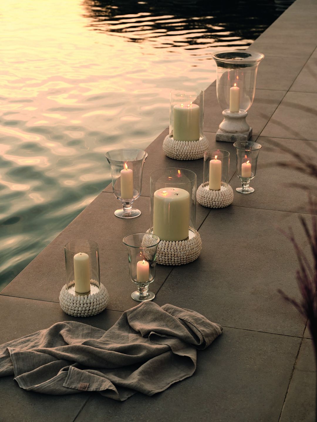 Several lit candles in glass holders sit on a stone surface near water, creating a warm and inviting atmosphere for outdoor decor.