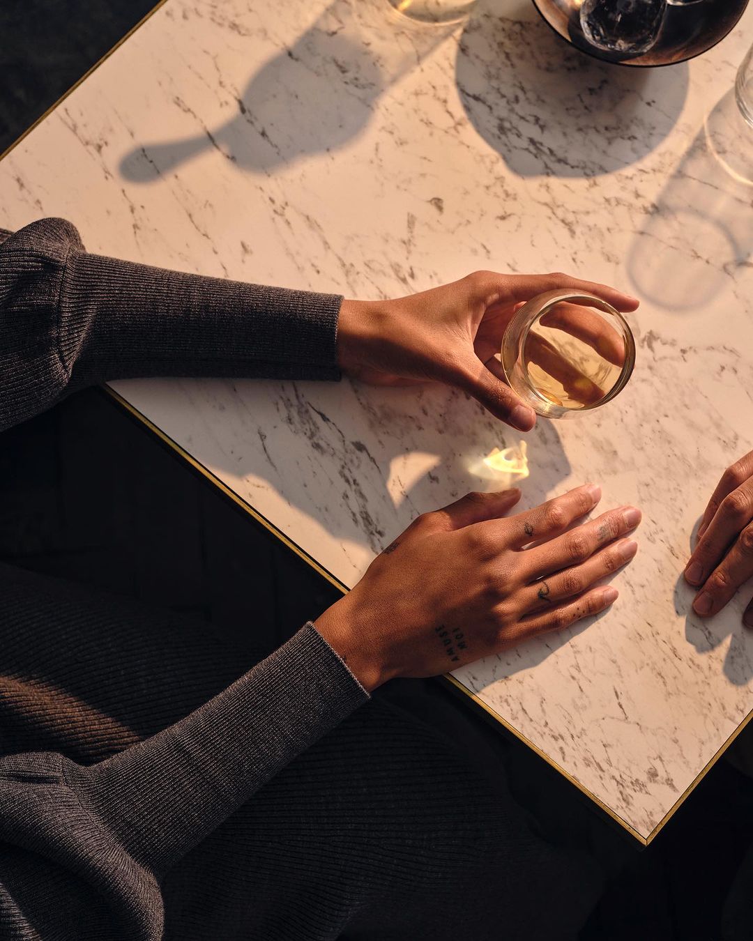 Overhead shot of two adult hands holding a glass of amber liquid on a marble table. The scene is softly lit, creating a warm ambiance.