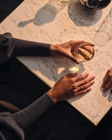 Overhead shot of two adult hands holding a glass of amber liquid on a marble table. The scene is softly lit, creating a warm ambiance.