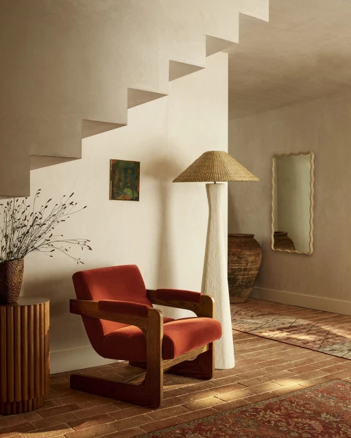 Interior view of a terracotta-coloured velvet chair, straw floor lamp, neutral wall with staggered steps and framed art.