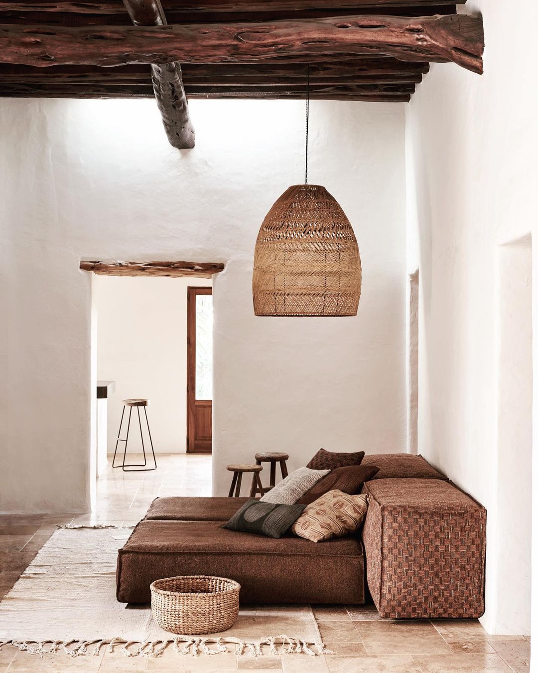 Brown woven pendant light and brown modular sofa with pillows, stools, and baskets on a stone floor in a minimalist interior space.