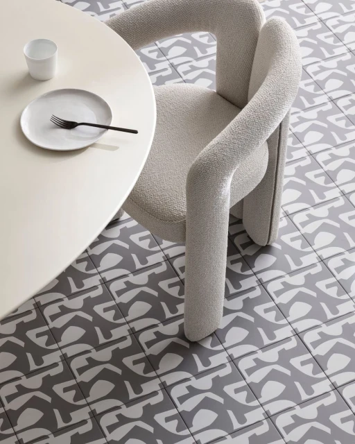 Interior scene featuring a modern chair with curved arms and a round table set with a plate, fork, and cup, set against a patterned tile floor.