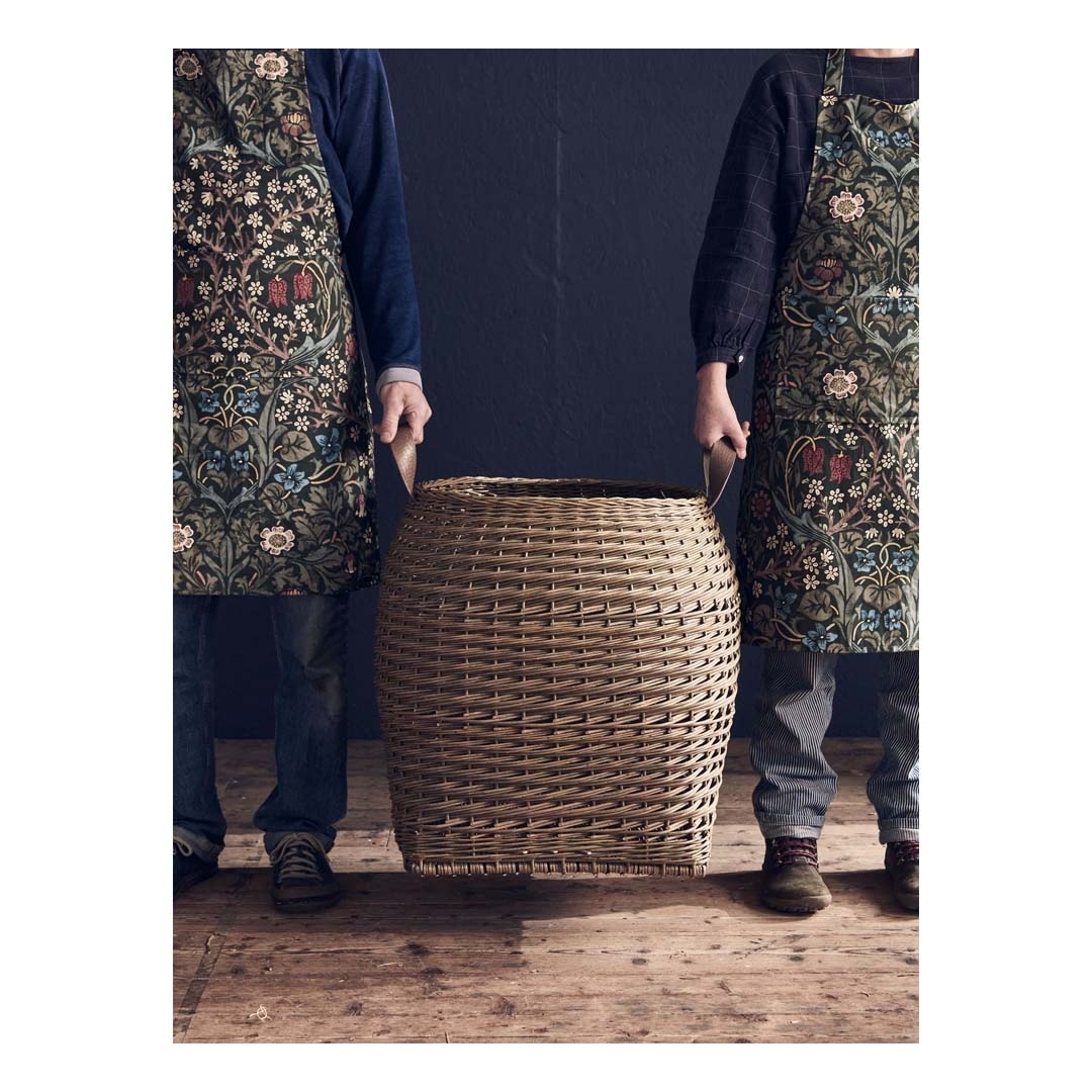 Two figures in floral aprons carry a woven laundry basket on a wood floor against a dark background, creating a vintage mood.