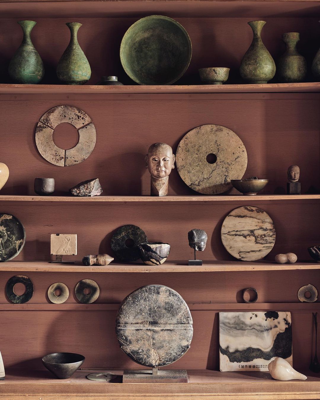 An assortment of pottery, ceramics, and stoneware are artfully arranged on multi-tiered shelves against a textured, reddish-brown backdrop.