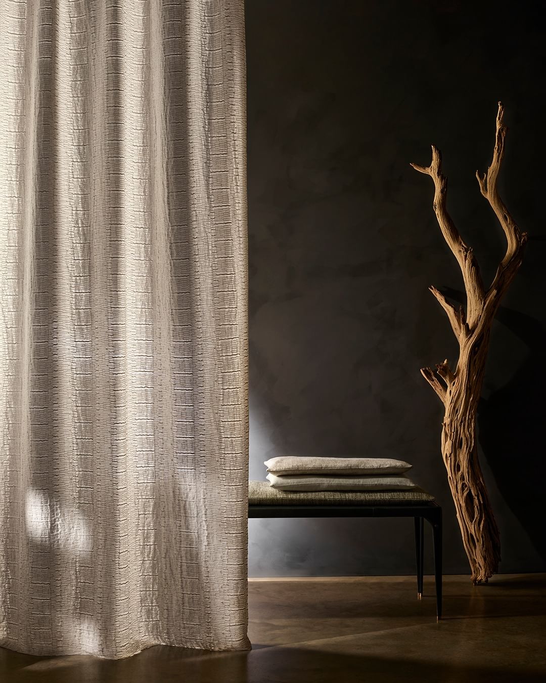 Textured curtain panel in a neutral tone with natural driftwood sculpture, and a small bench with cushions against a dark wall.