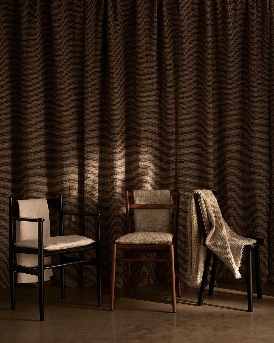 Interior design shot featuring three empty wooden chairs arranged in front of textured brown curtains, creating a minimalist aesthetic.