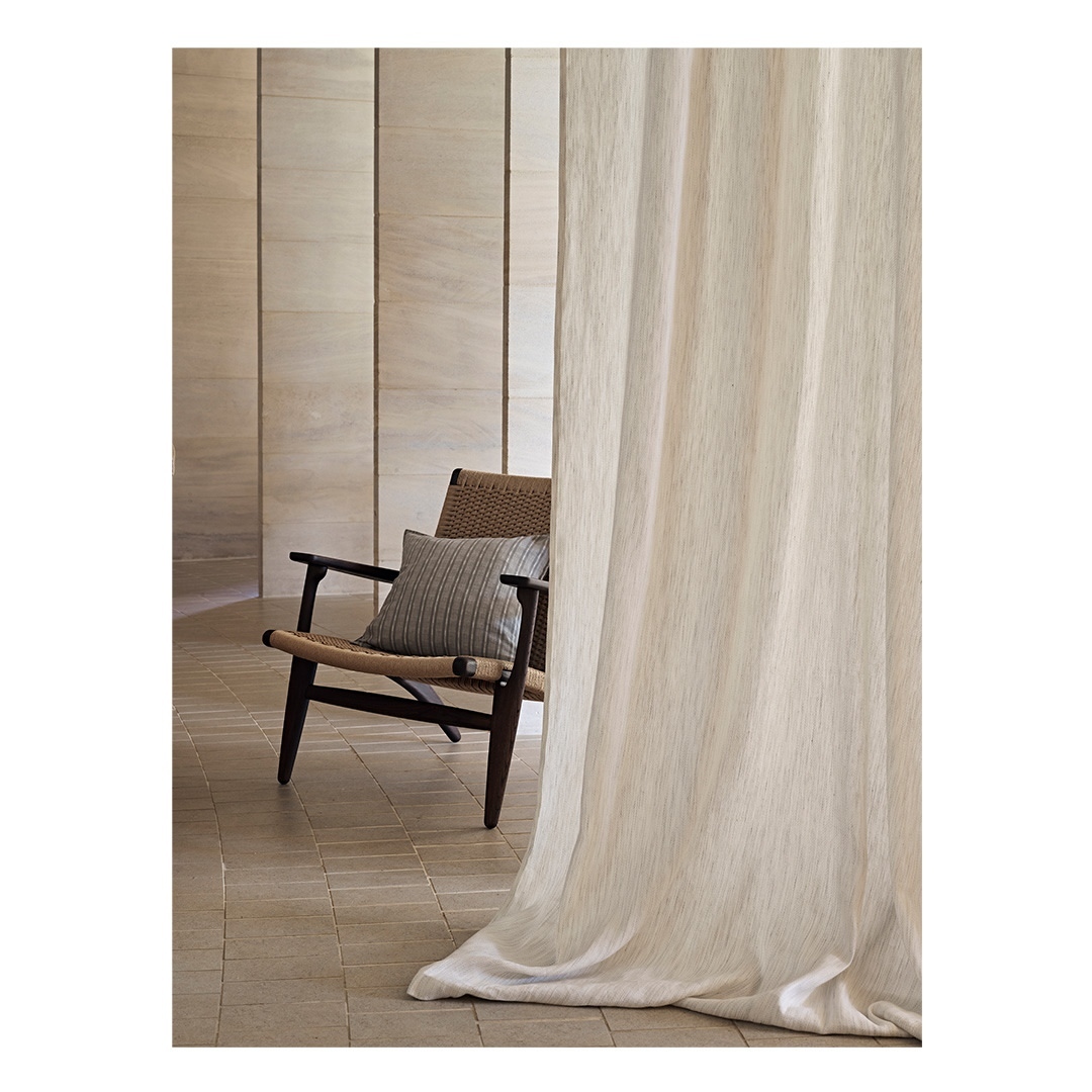 Interior view of a woven chair with a striped cushion next to a light-colored curtain in front of a vertically paneled wall.