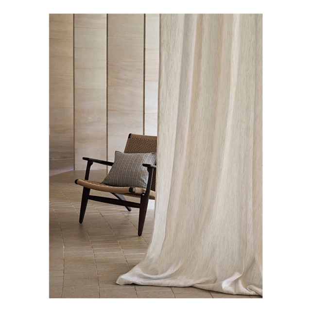 Interior view of a woven chair with a striped cushion next to a light-colored curtain in front of a vertically paneled wall.