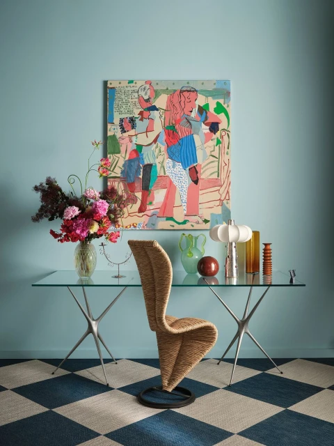 Living room interior with glass-top table, woven chair, vibrant artwork, and floral arrangement against a blue wall and checkered rug.