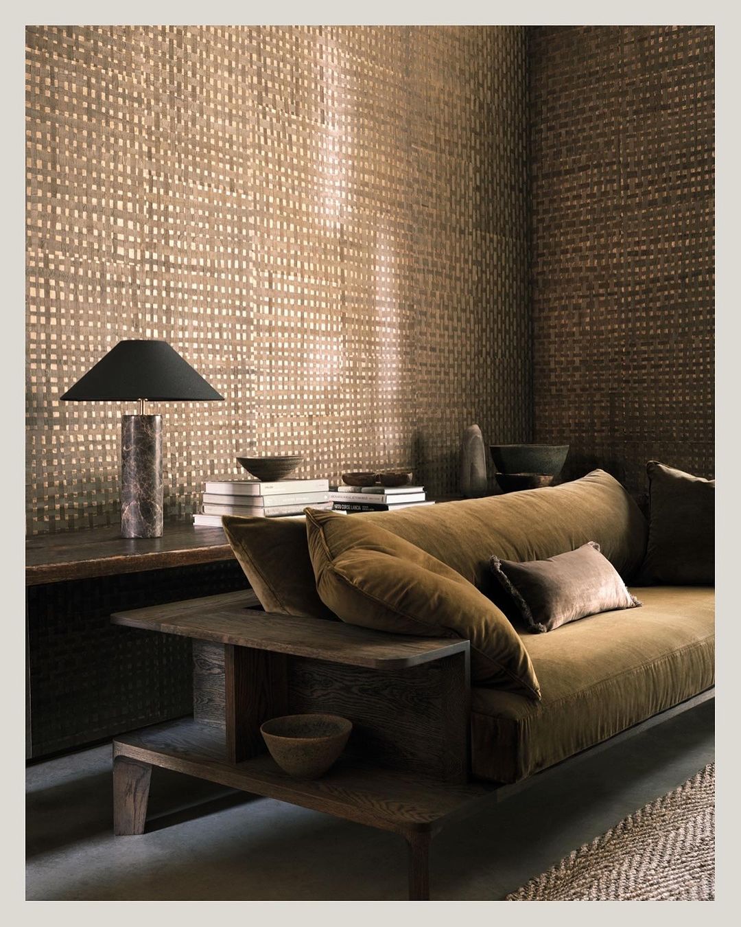Living room interior with a sofa, a lamp on a side table with books, and a woven textured wall covering in neutral brown tones.