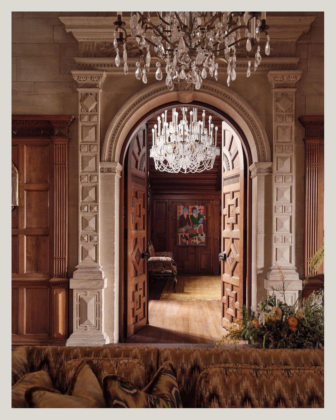 Ornate interior with open carved wood doors, crystal chandeliers, and a tufted brown couch in the foreground. Elegant design.