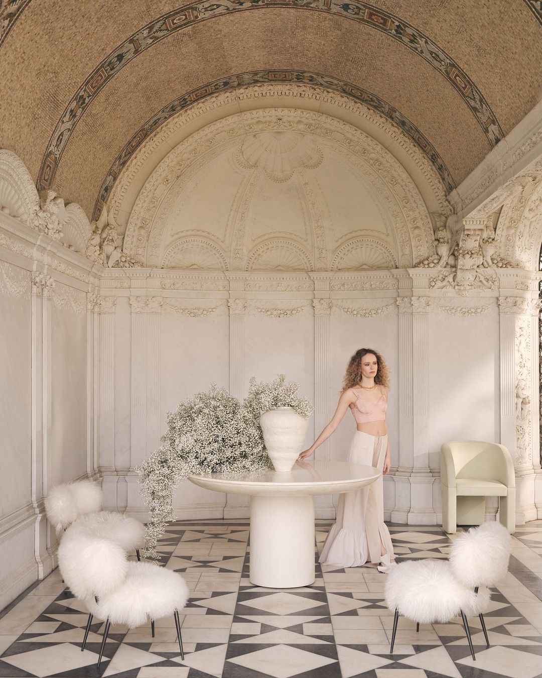 Indoor shot of a curly-haired adult woman wearing a pink top and white skirt next to a table with a vase of baby's breath flowers.