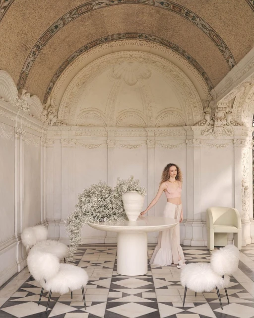 Indoor shot of a curly-haired adult woman wearing a pink top and white skirt next to a table with a vase of baby's breath flowers.