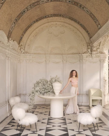 Indoor shot of a curly-haired adult woman wearing a pink top and white skirt next to a table with a vase of baby's breath flowers.