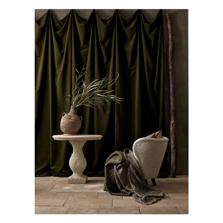 Interior setting with a green velvet curtain backdrop, table with vase, and neutral-toned armchair. Cozy, elegant indoor composition.