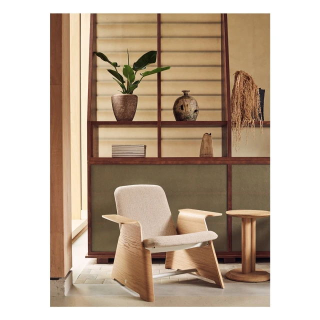 Beige upholstered armchair with light wood frame in a neutral indoor setting with a shelving unit and side table.