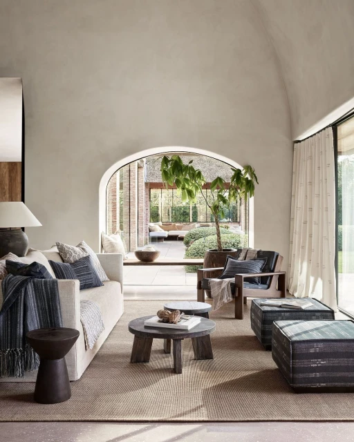 Living room interior with a white sofa, wooden chairs, coffee tables, and a large window offering a view of an outdoor area.