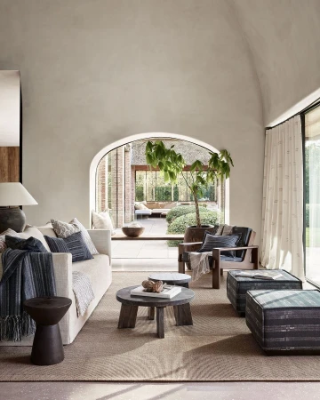 Living room interior with a white sofa, wooden chairs, coffee tables, and a large window offering a view of an outdoor area.