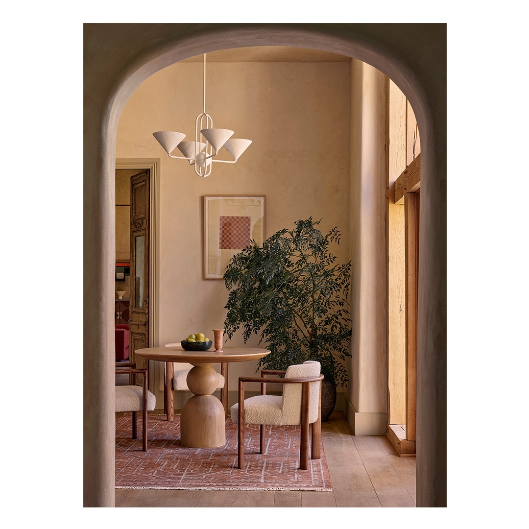 Interior view of a modern dining room with a round wooden table, upholstered chairs, chandelier lighting, and a large potted plant.