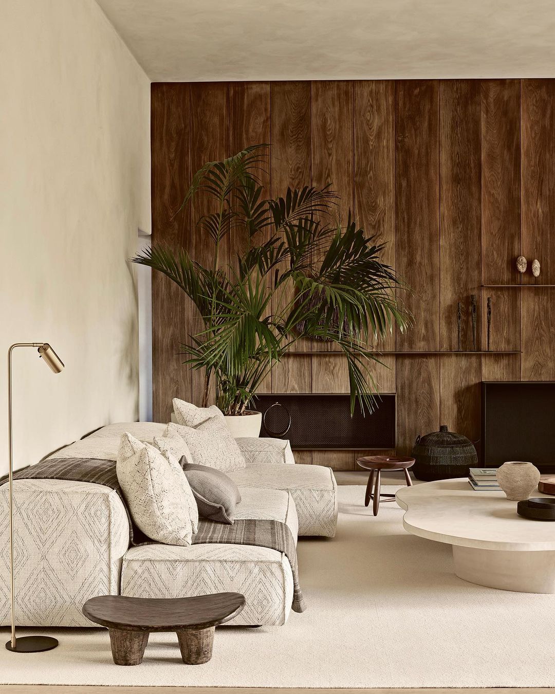 Living room interior featuring a patterned sofa, light rug, dark wood accent wall, potted plant, and neutral tones in a modern setting.