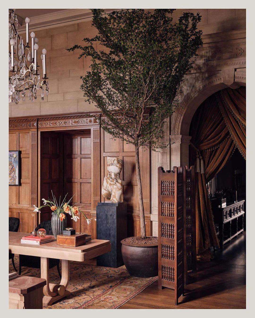 Opulent interior with wooden panelling, chandelier, table with floral arrangement, potted tree, and decorative screen in warm tones.