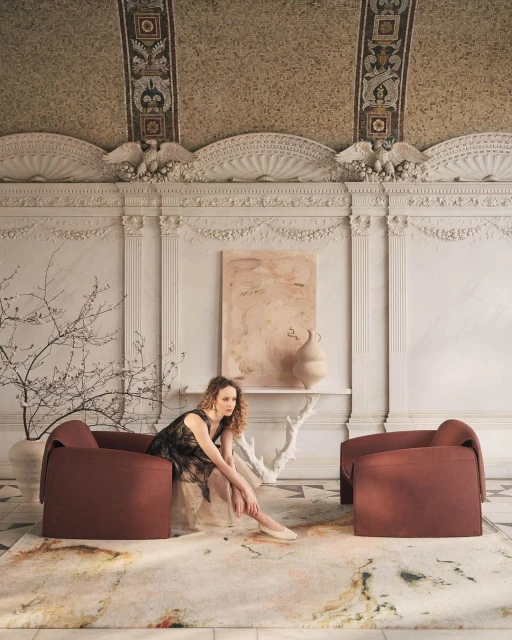 A woman in a black dress sits in a rust-colored armchair on a patterned rug, in a room with ornate plasterwork and modern art pieces.