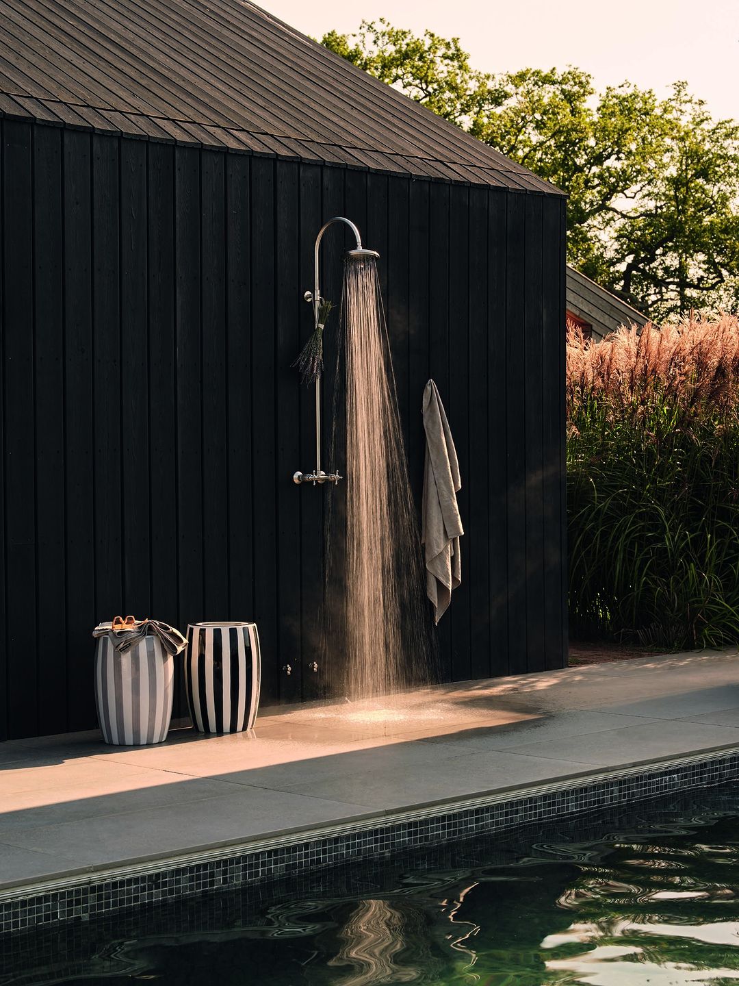 Outdoor shower with running water next to a pool, featuring striped stools and a dark wood background under natural daylight.