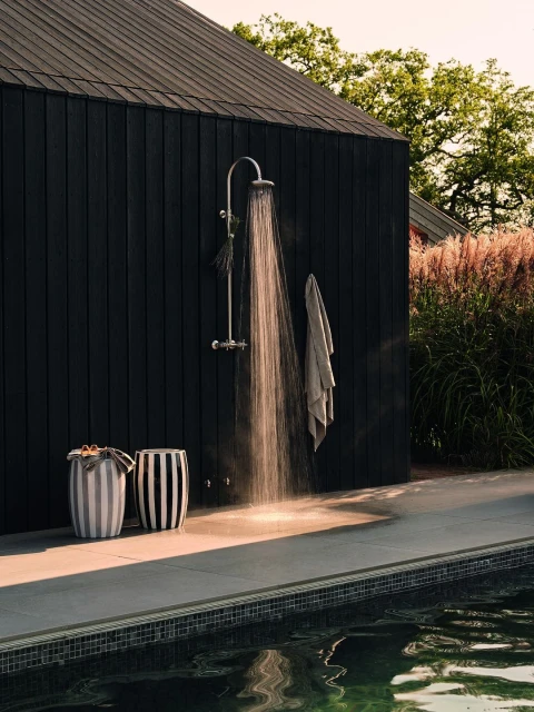 Outdoor shower with running water next to a pool, featuring striped stools and a dark wood background under natural daylight.
