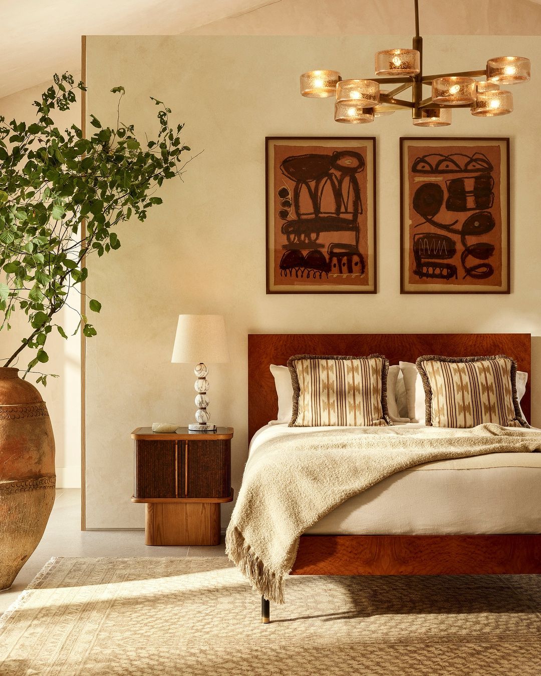 Bedroom interior featuring a bed with neutral linens, decorative pillows, wall art, a side table, table lamp, and a large plant in a vase.