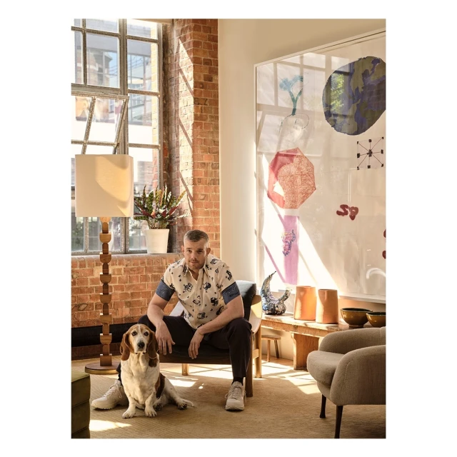 Adult male sitting indoors with his dog, wearing a patterned shirt, black pants, and sneakers, posing near a floor lamp and large window.