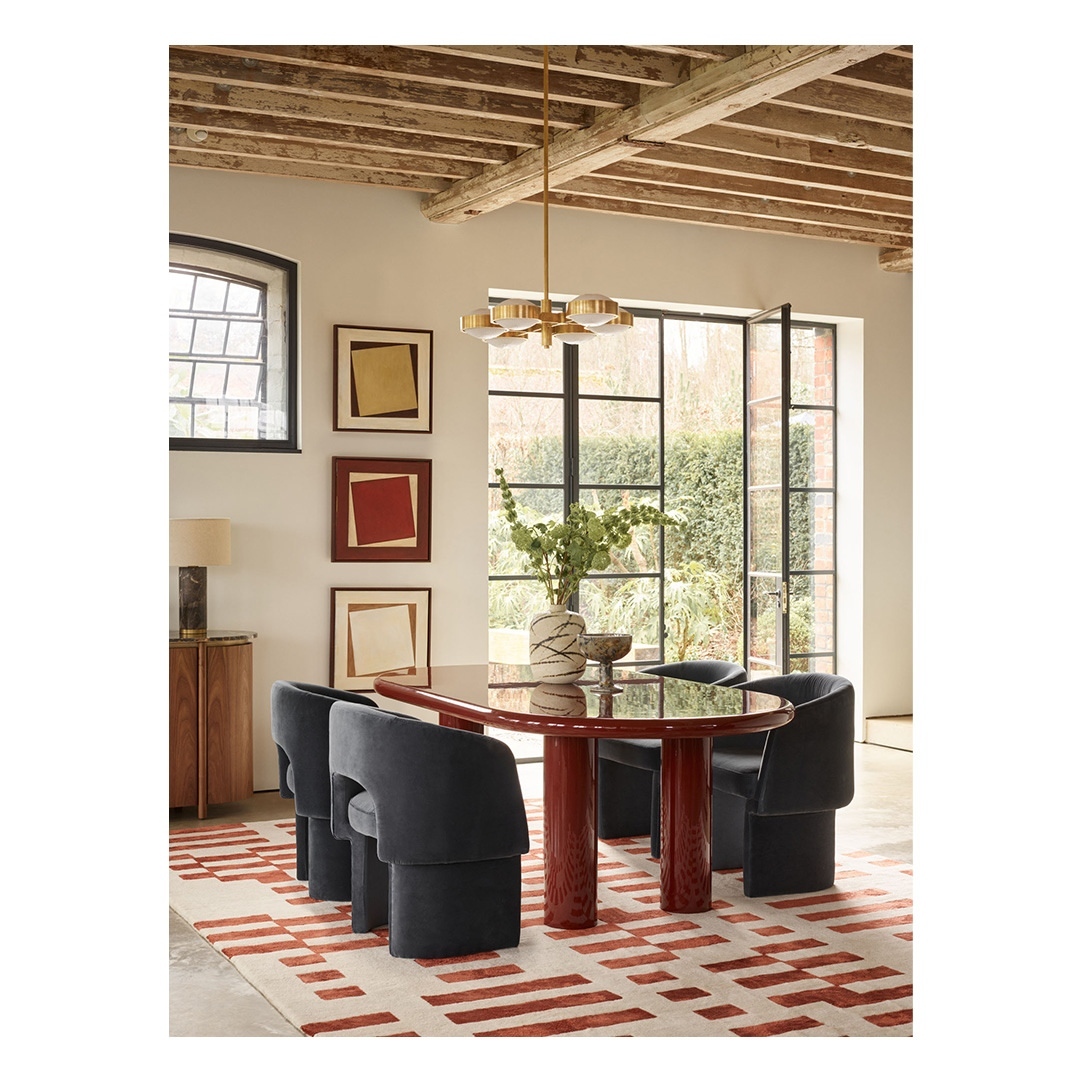 Contemporary dining room with red oval table, blue velvet chairs, and patterned rug, illuminated by modern chandelier.