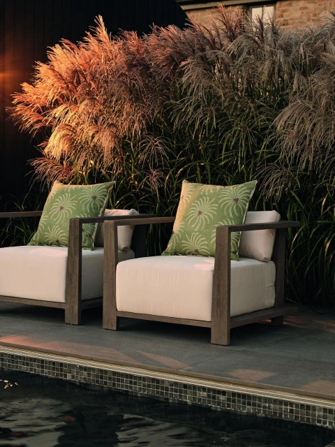 Outdoor lounge setting features two armchairs with light cushions and green pillows, complemented by ornamental grass in the background.