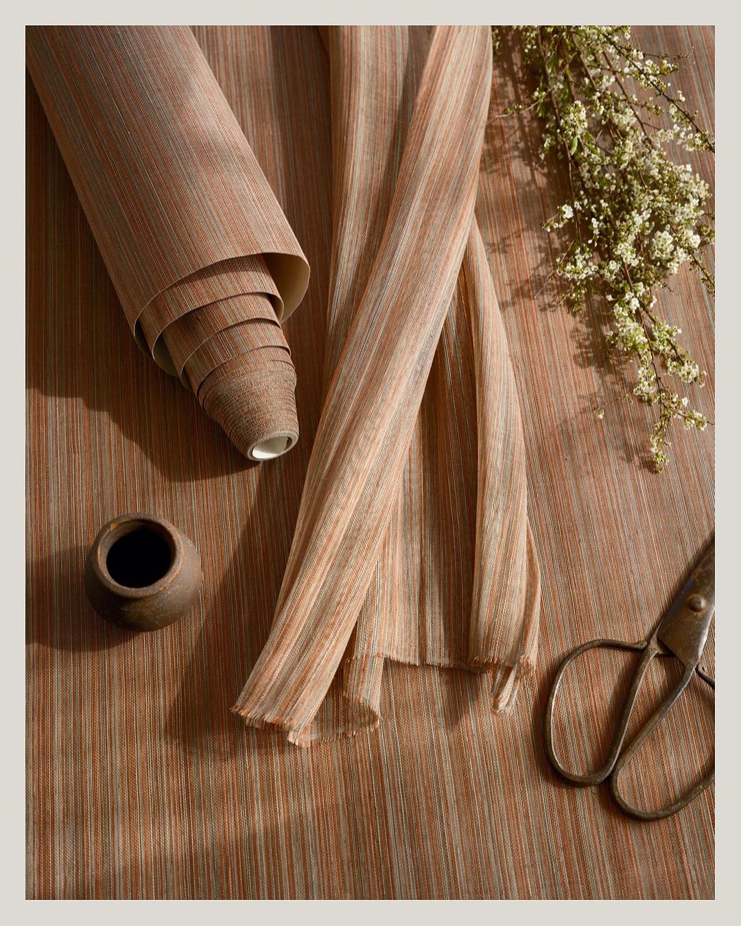 Roll of textured wallpaper alongside cut lengths, styled with ceramic vase, antique scissors, and sprigs of white flowers on wood-grain surface.