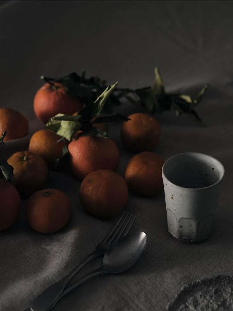 Still life photograph of oranges, a ceramic cup, and silverware arranged on a textured linen surface in dim, moody lighting.