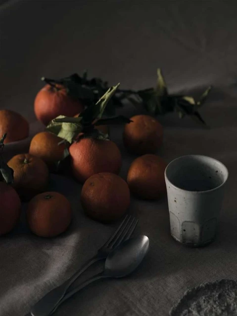 Still life photograph of oranges, a ceramic cup, and silverware arranged on a textured linen surface in dim, moody lighting.