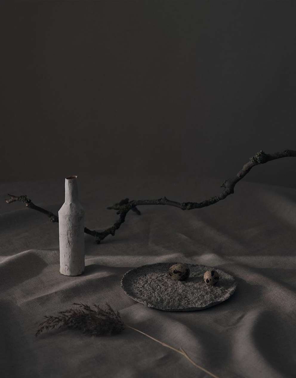 Still life photography of a bottle, two quail eggs on a plate, and dried grass against a mottled grey background with soft studio lighting.