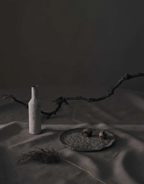 Still life photography of a bottle, two quail eggs on a plate, and dried grass against a mottled grey background with soft studio lighting.