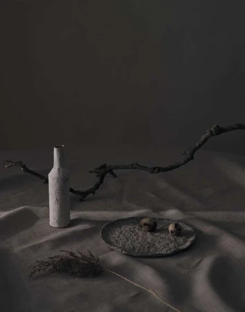Still life photography of a bottle, two quail eggs on a plate, and dried grass against a mottled grey background with soft studio lighting.