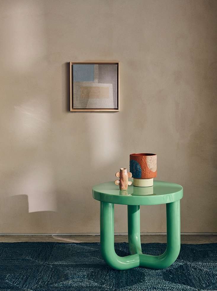 Interior design featuring a green round table with decor objects, a colorful vase, and framed art against a neutral-toned wall. 