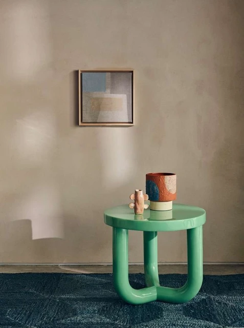 Interior design featuring a green round table with decor objects, a colorful vase, and framed art against a neutral-toned wall. 