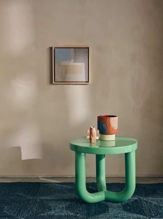 Interior design featuring a green round table with decor objects, a colorful vase, and framed art against a neutral-toned wall. 
