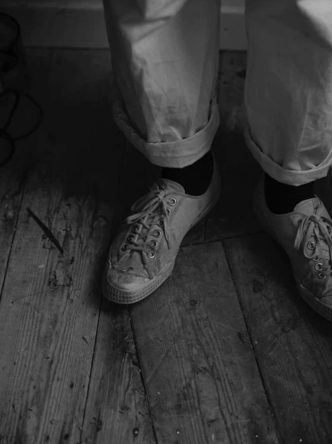 Black and white image featuring a pair of worn canvas sneakers, light-colored rolled pants, and a weathered wood floor.