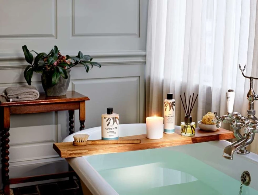 Bathroom with Cowshed bath products, lit candle, and natural sponge on a wooden tray over the white porcelain bathtub indoors.