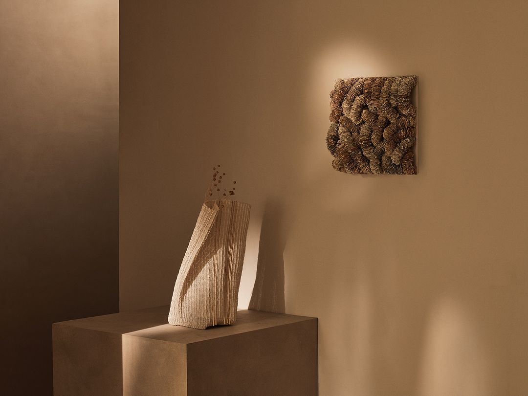 Beige toned still life with a textured vase on a plinth and a square textured wall hanging, soft lighting and neutral color palette.