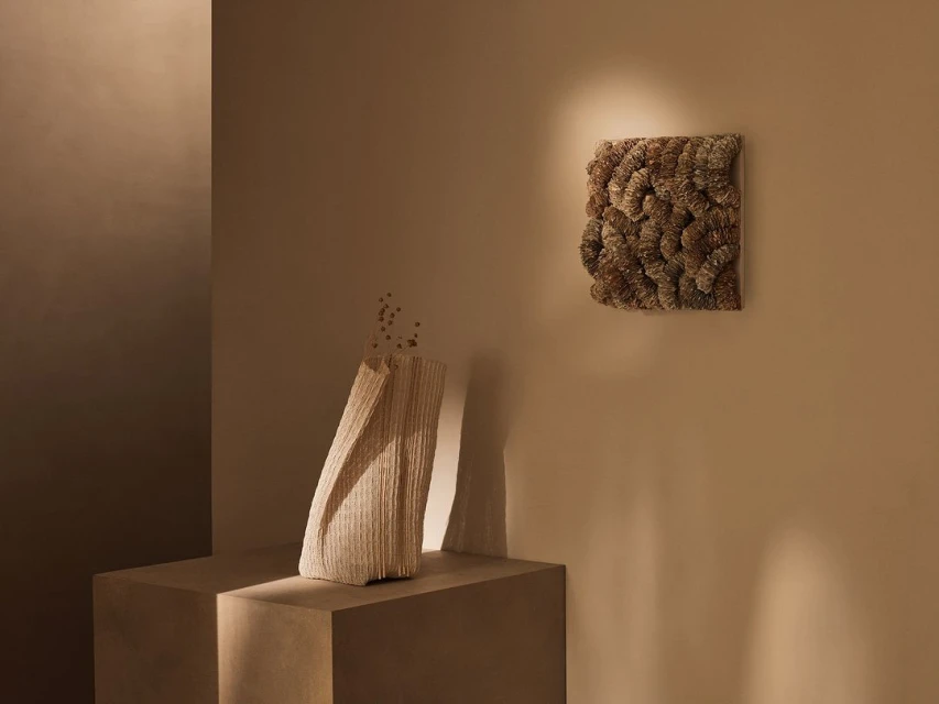 Beige toned still life with a textured vase on a plinth and a square textured wall hanging, soft lighting and neutral color palette.