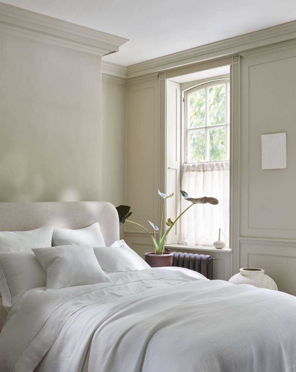 Bedroom interior with a white linen-covered bed, a radiator, and decor, bathed in neutral tones and soft natural light from the window.